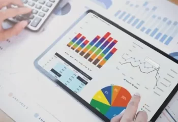 A imagem mostra uma mão usando um tablet com gráficos e dados visuais sobre uma mesa, ao lado de uma calculadora, ideal para análise de métricas de vendas.