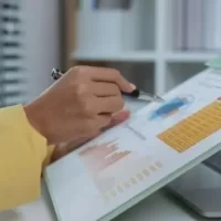 Imagem de uma pessoa analisando gráficos e dados em um caderno enquanto utiliza um laptop ao fundo, representando a importância da análise de dados para tomada de decisões