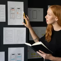 Designers analisando protótipos de aplicativos móveis para desenvolvimento de produto em uma parede com sketches e wireframes.