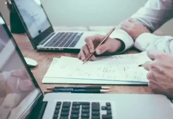 Duas pessoas trabalhando em uma mesa com laptops e anotações, discutindo um projeto juntos