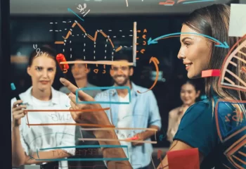 Equipe de profissionais discutindo estratégias de marketing digital, com gráficos e anotações em uma tela de vidro, demonstrando planejamento e inovação.