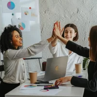 Três mulheres de escritório comemorando uma conquista, símbolo de sucesso em estratégias de go-to-market, em ambiente de trabalho colaborativo com gráficos.