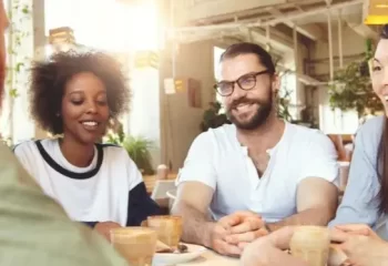 Grupo de amigos sorrindo e conversando em um café, rodeados por plantas. A imagem retrata um ambiente descontraído e social.