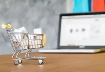Miniatura de carrinho de supermercado sobre mesa de madeira com notebook ao fundo