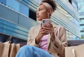 Mulher estilosa com um smartphone, sentada em um banco, cercada por sacolas de compras em ambiente urbano moderno.