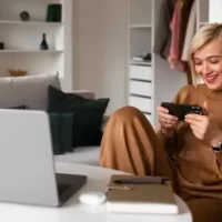 Mulher sorridente jogando no smartphone enquanto está sentada em um ambiente confortável com um laptop e itens de trabalho ao redor.