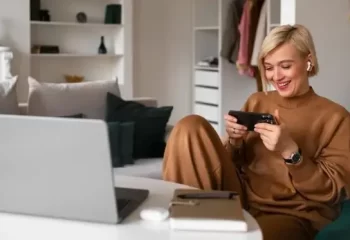 Mulher sorridente jogando no smartphone enquanto está sentada em um ambiente confortável com um laptop e itens de trabalho ao redor.