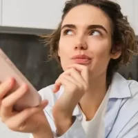 Mulher pensativa segurando um smartphone em uma cozinha moderna, refletindo sobre suas opções.