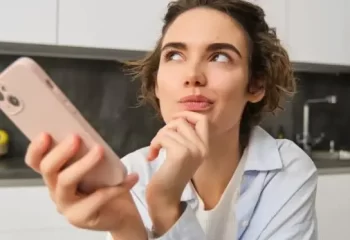 Mulher pensativa segurando um smartphone em uma cozinha moderna, refletindo sobre suas opções.