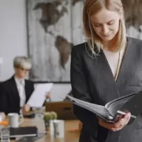 Uma mulher em trajes formais revisando documentos em uma sala de reuniões, com mapas ao fundo e outras pessoas trabalhando.
