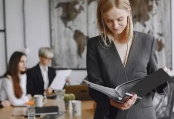 Uma mulher em trajes formais revisando documentos em uma sala de reuniões, com mapas ao fundo e outras pessoas trabalhando.