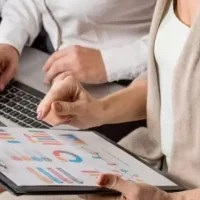 Mulher sentada apontando para gráficos em prancheta nas mãos, junto à colega de trabalho com notebook no colo