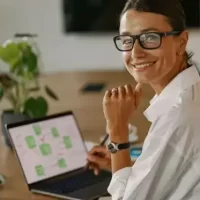 Mulher sorridente usando óculos em ambiente de trabalho moderno, olhando para a câmera enquanto trabalha em laptop sobre mesa de madeira.