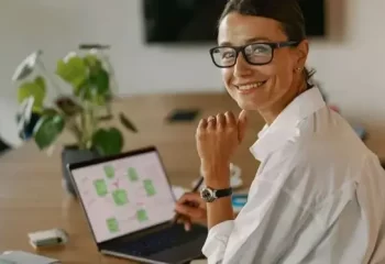 Mulher sorridente usando óculos em ambiente de trabalho moderno, olhando para a câmera enquanto trabalha em laptop sobre mesa de madeira.
