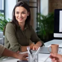 Mulher sorrindo em uma reunião no escritório com colegas, enquanto uma videoconferência ocorre em um monitor ao fundo.