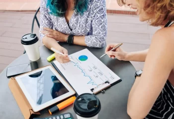 Duas mulheres analisando gráficos financeiros e dados em papel com tablet, smartphone e café em mesa redonda