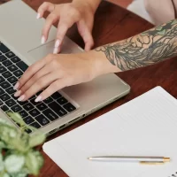 Pessoa trabalhando em um laptop com um braço tatuado em uma mesa de madeira, com caderno e caneta ao lado, em ambiente de escritório ou estudo.