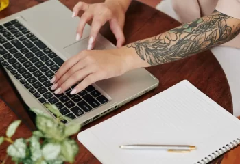 Pessoa trabalhando em um laptop com um braço tatuado em uma mesa de madeira, com caderno e caneta ao lado, em ambiente de escritório ou estudo.