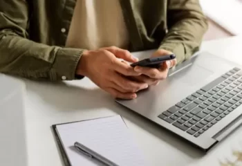 Pessoa usando smartphone, com notebook e bloco de anotações sobre a mesa