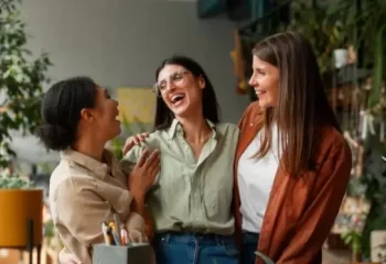 Três mulheres sorrindo e se divertindo em um ambiente leve e descontraído, cercadas por plantas e decoração moderna.